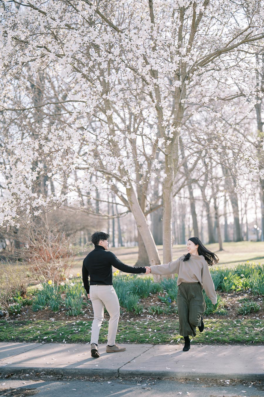 A joyful couple runs hand in hand beneath a blooming cherry blossom tree in a park, capturing a moment of happiness and springtime beauty.