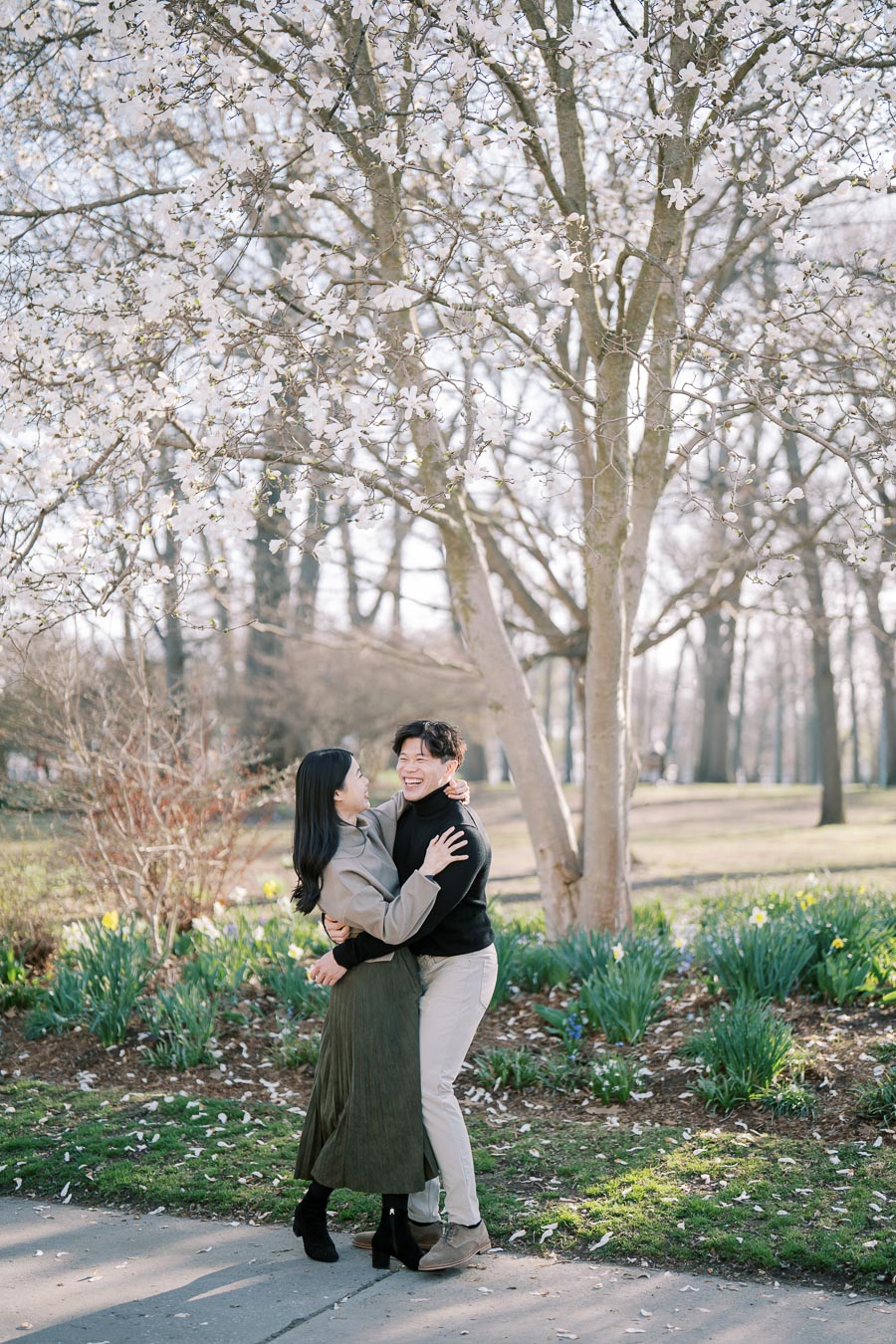 A couple shares a joyful embrace under a blooming tree in a park, surrounded by spring flowers and greenery.