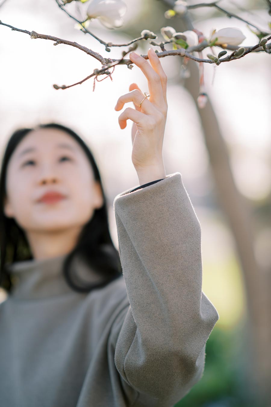 A person in a gray coat gently reaches towards a budding branch, capturing a moment of connection with nature.