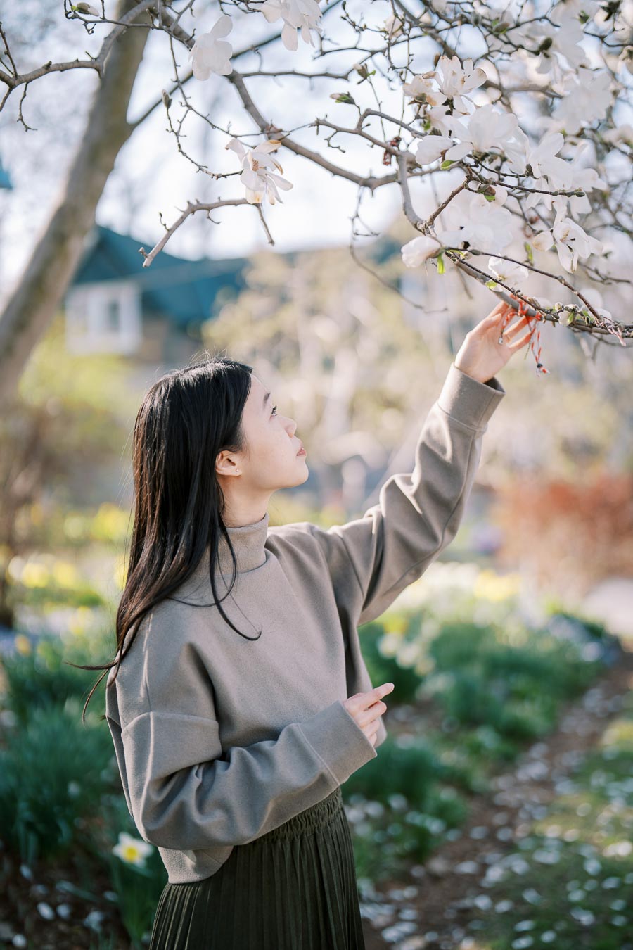 A woman gently touches a branch of a blossoming tree in a garden, surrounded by spring flowers.
