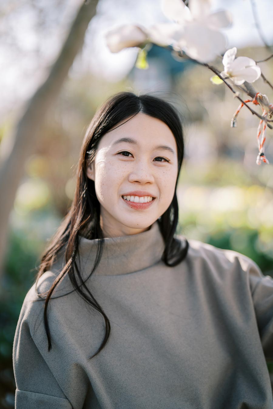 A woman smiling outdoors with a blurred background of trees and flowers, wearing a gray turtleneck sweater.