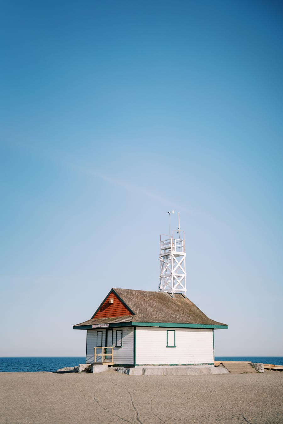 A small beachside building with a tower stands on a sandy shore, overlooking the ocean under a clear blue sky.