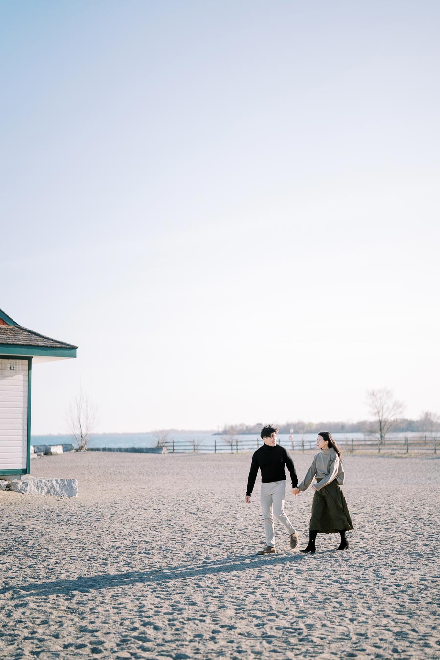A couple strolls hand in hand along a sandy beach, with a building and a serene body of water in the background.