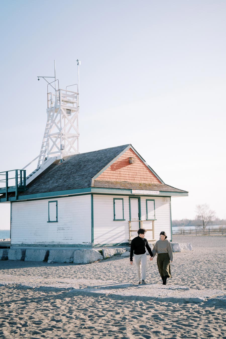 A couple walks hand in hand on a sandy beach near a small white building with a tower, under a clear blue sky.