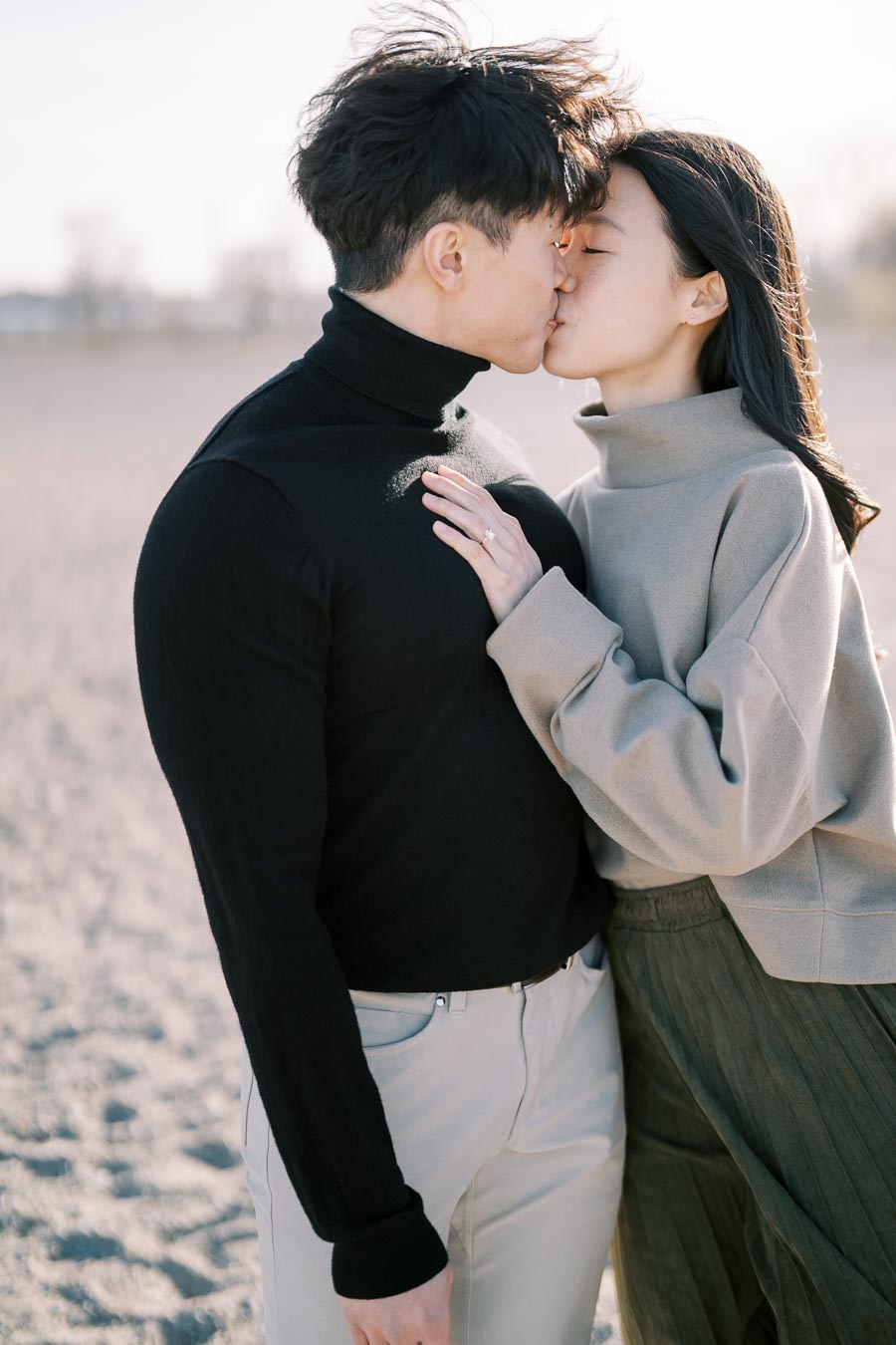 A couple shares a kiss outdoors, dressed in casual attire with a sandy background.