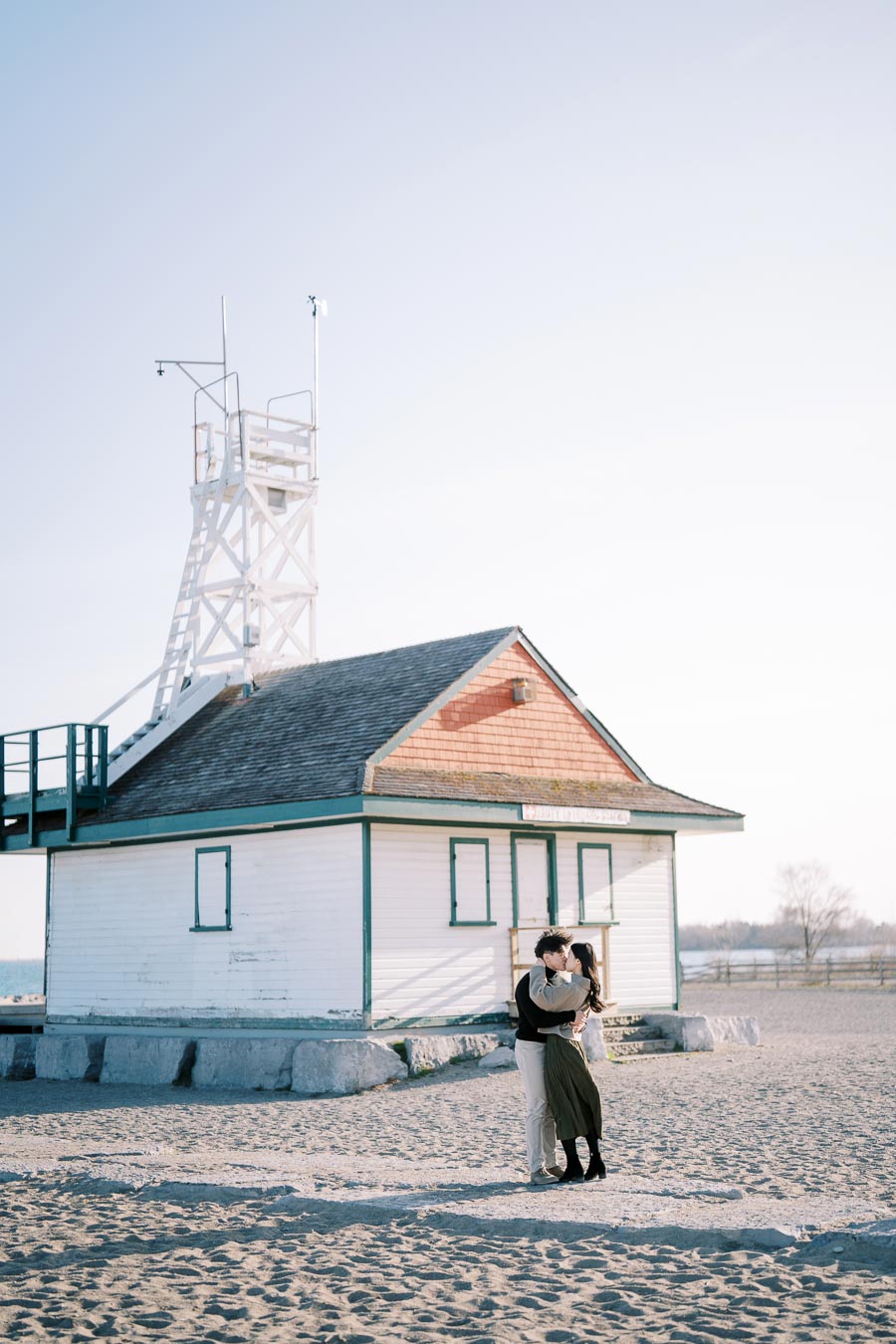A couple shares a tender moment in front of a quaint beach building with a tower, set against a clear sky.