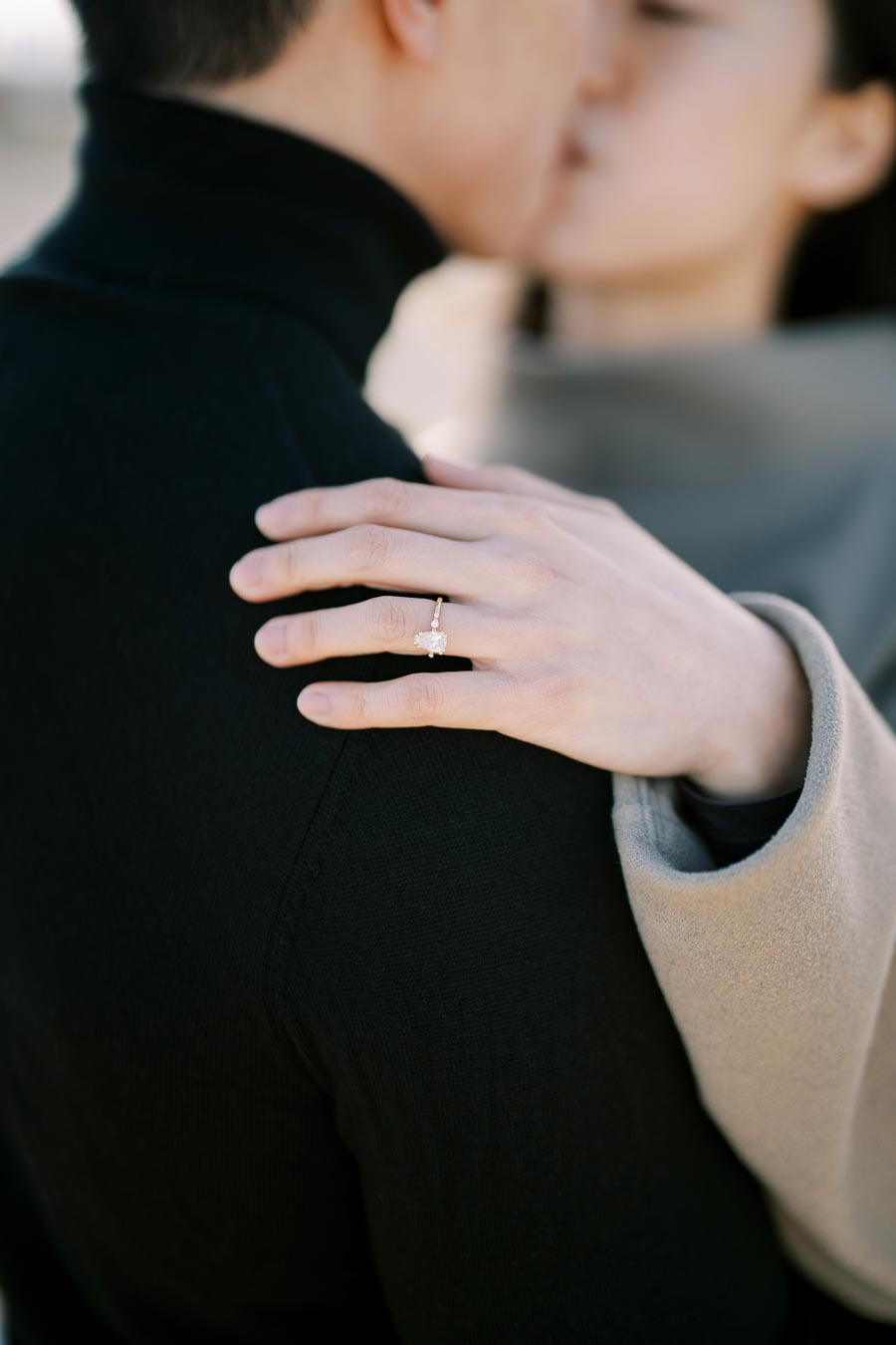 A close-up shot of a couple embracing, highlighting a woman's hand with an engagement ring resting on the man's shoulder.