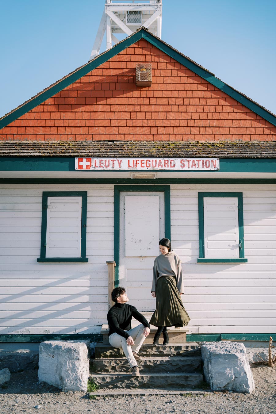 A couple poses in front of the historic Leuty Lifeguard Station, showcasing its distinctive red shingles and green trim.