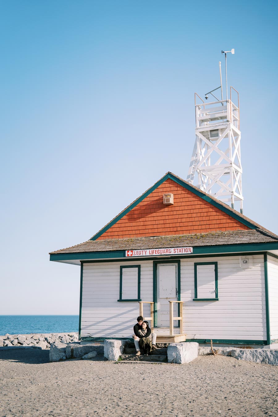 A couple sits in front of the Leuty Lifeguard Station, a historic building located by the beach, under a clear blue sky.