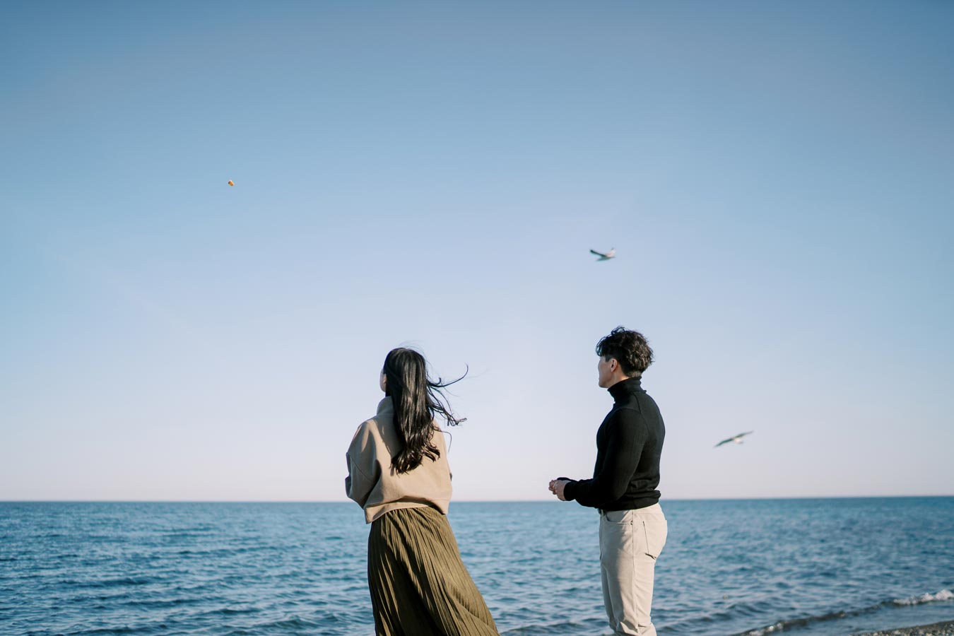 A couple stands on a beach, gazing at the sea and sky as birds fly overhead, creating a serene and peaceful scene.
