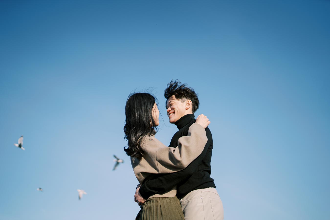 A couple embraces under a clear blue sky, surrounded by flying seagulls, capturing a moment of joy and connection.