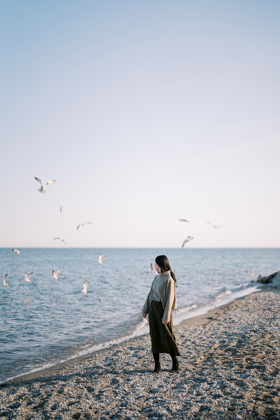 A person stands on a pebbly beach, gazing at seagulls flying over the calm sea under a clear sky.