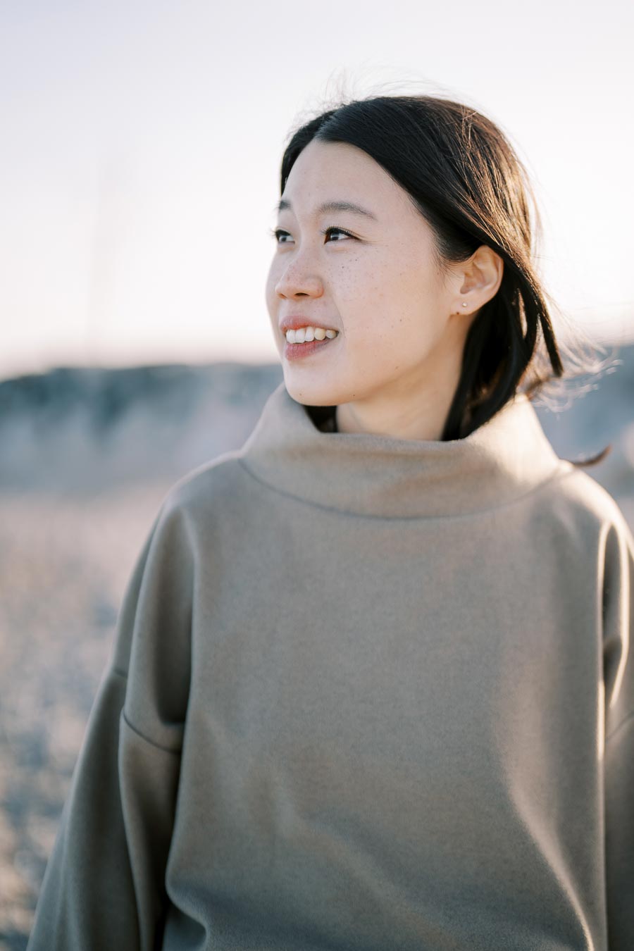 A woman smiles as she looks into the distance, wearing a beige sweater and standing outdoors.