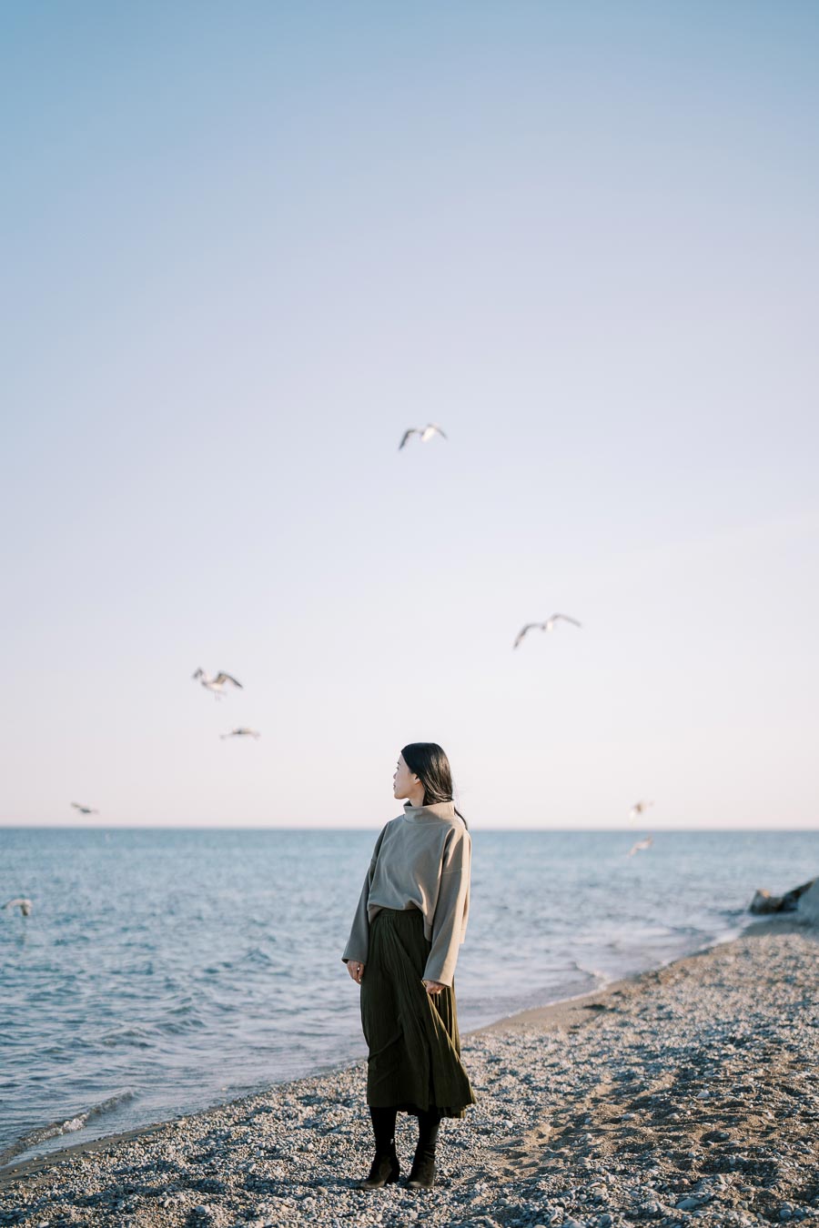 A person stands on a pebbled beach, gazing at the sea as seagulls fly overhead.