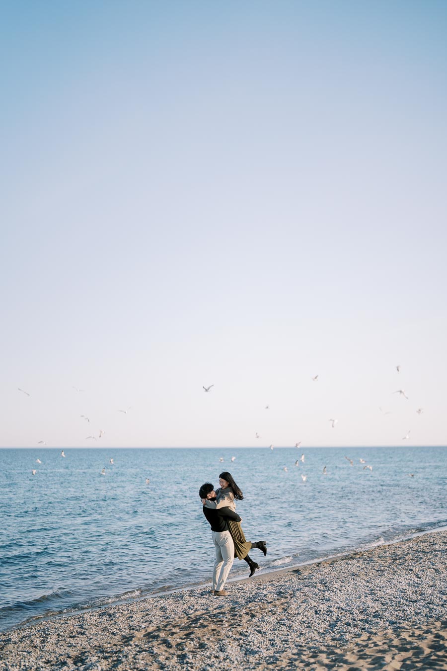A couple shares a joyful moment on a beach, surrounded by flying seagulls and the serene ocean.