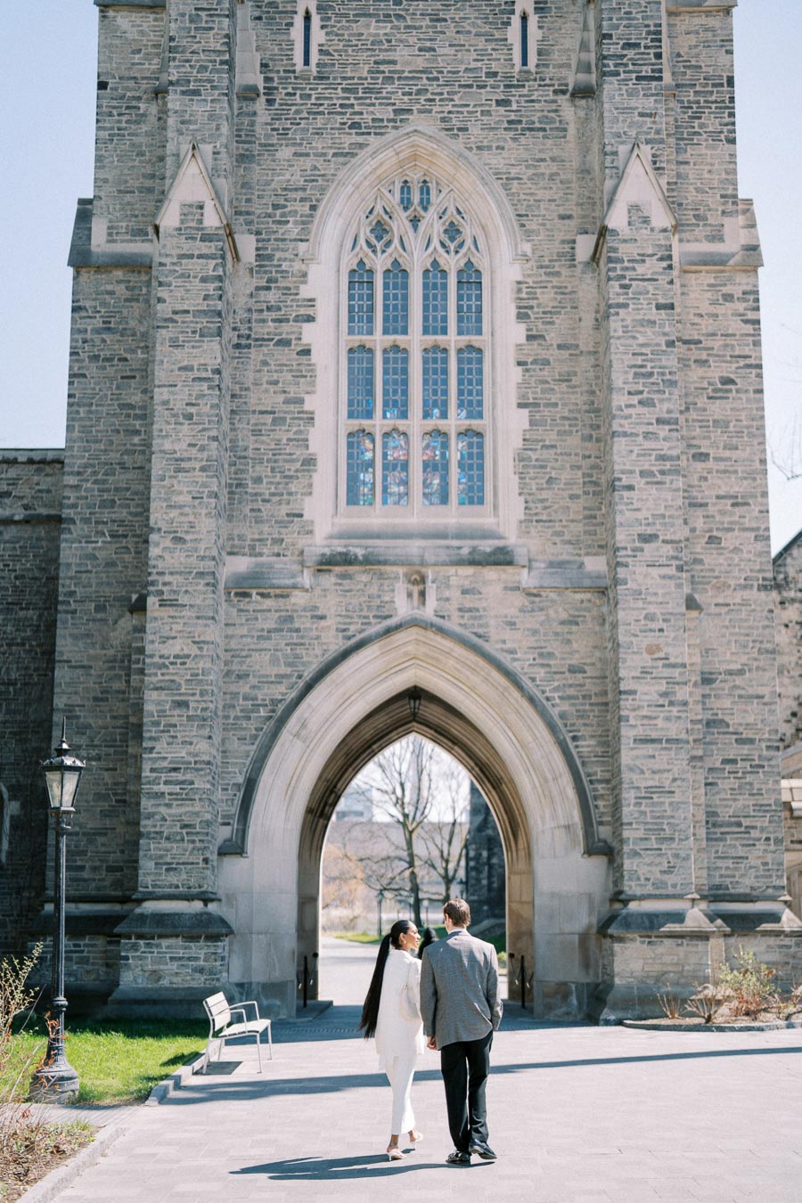 Couple walking hand in hand under a large stone archway at a university campus.