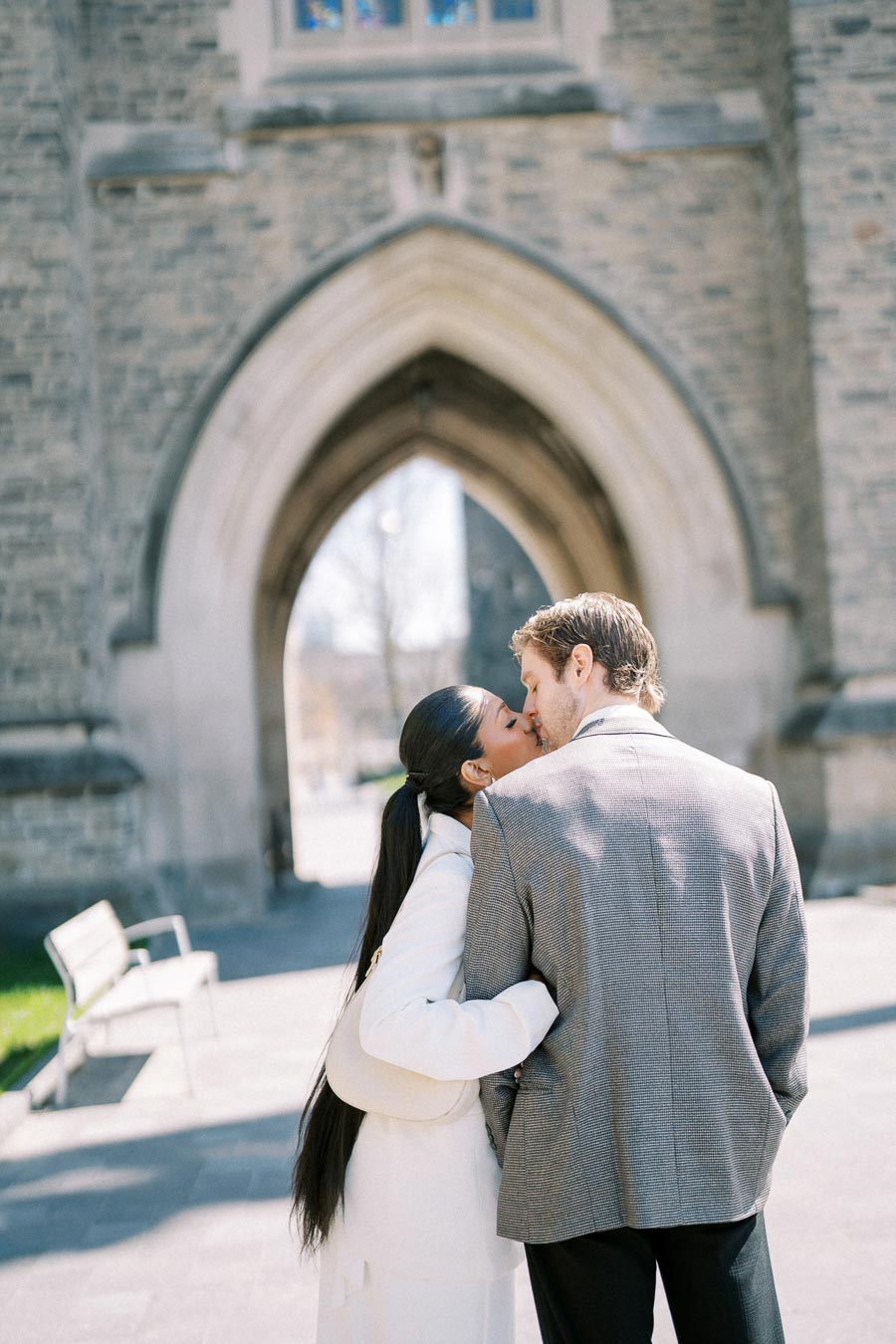 A couple kissing in front of a historic stone building with an arched entrance, wearing formal attire.