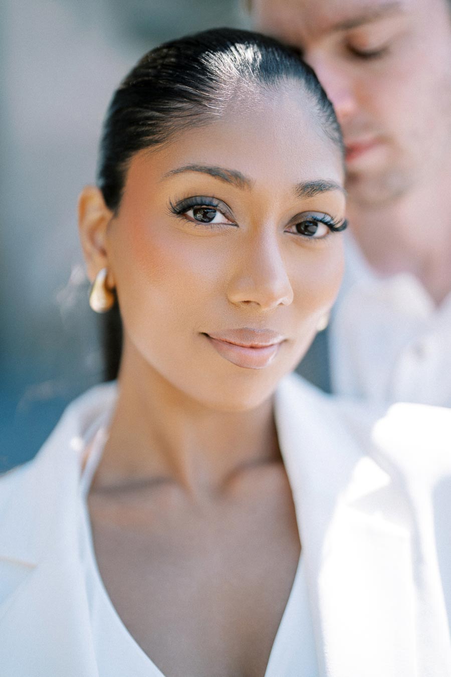 Close-up portrait of a woman with long hair and earrings, with a man partially visible in the background.