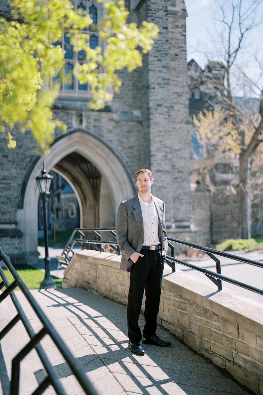 Man wearing a gray blazer and black pants standing near a stone archway at the University of Toronto.