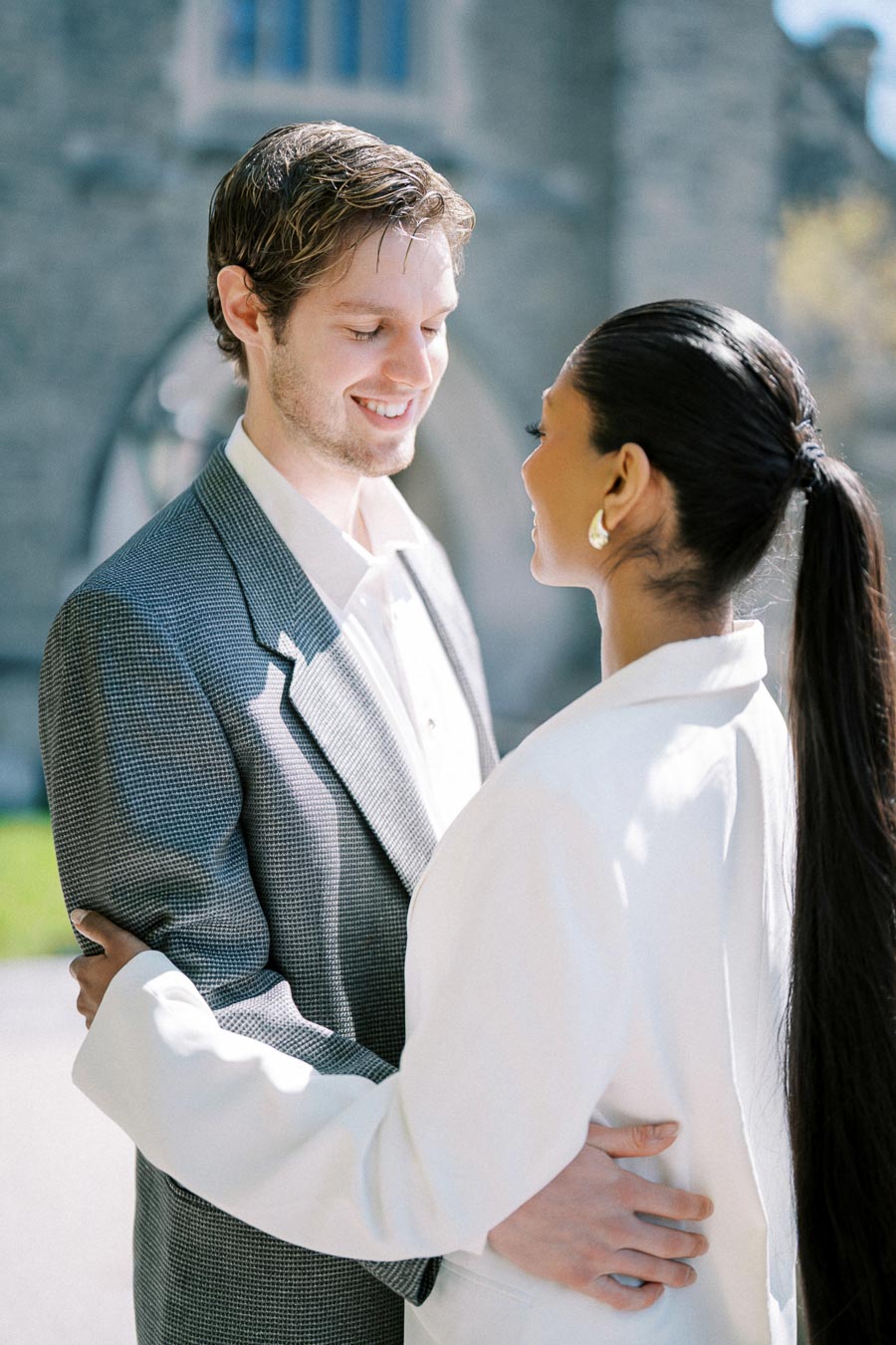 A couple embracing outdoors at the University of Toronto, with the woman wearing a white blazer and the man in a patterned suit.
