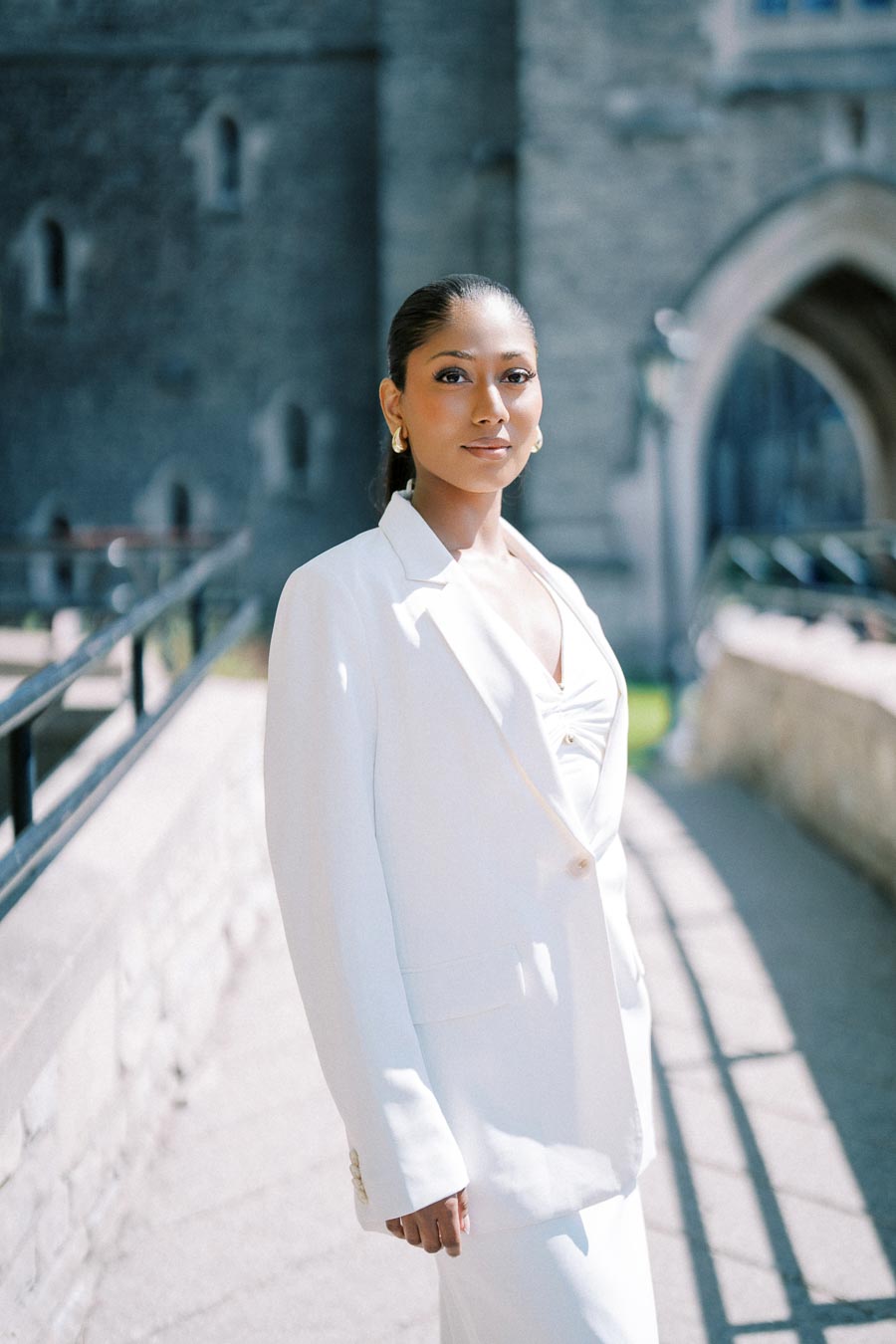 Elegant woman in a white suit standing outdoors in front of a historic stone building.