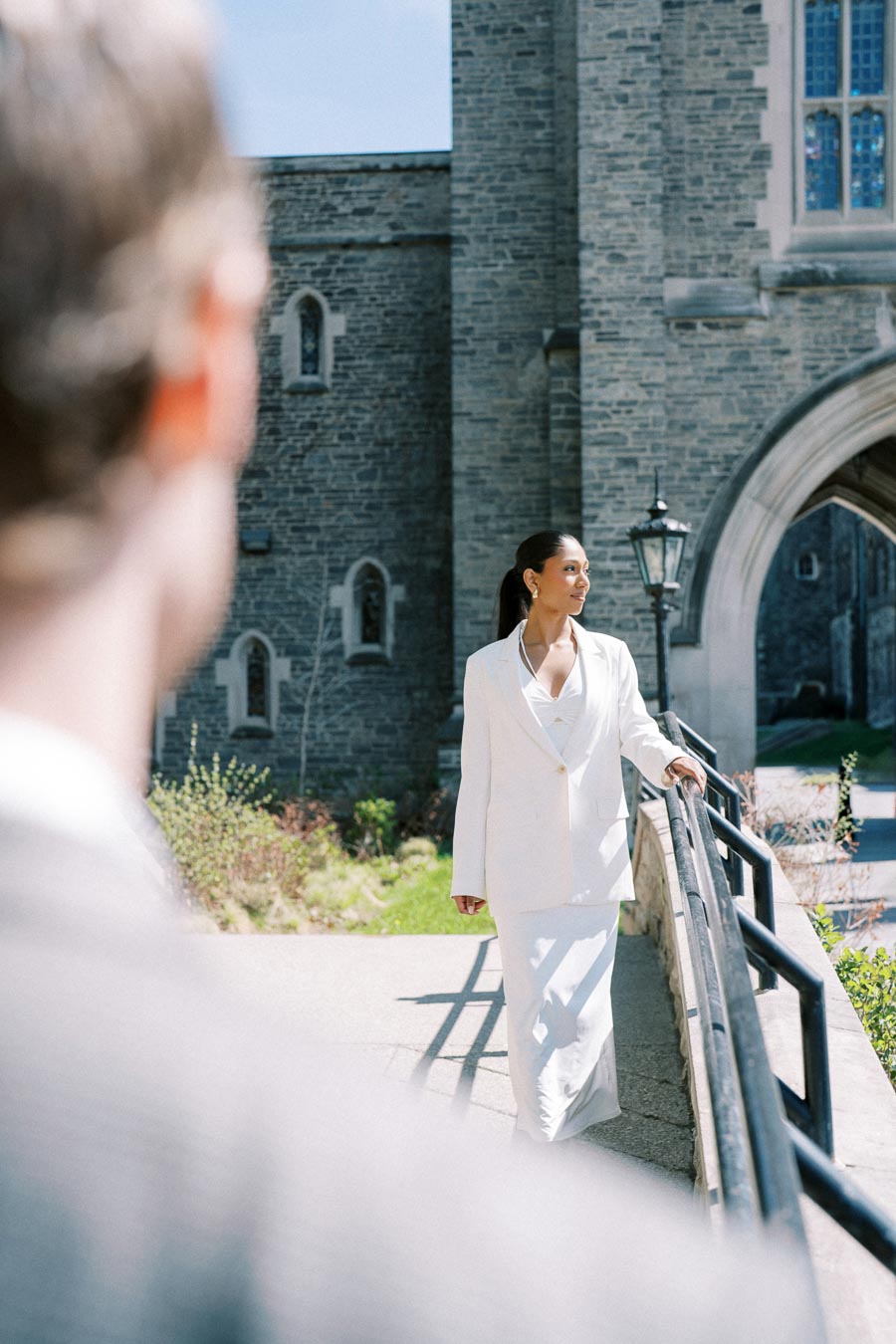 A woman in a white suit standing on a railing at the University of Toronto with a man in the background.