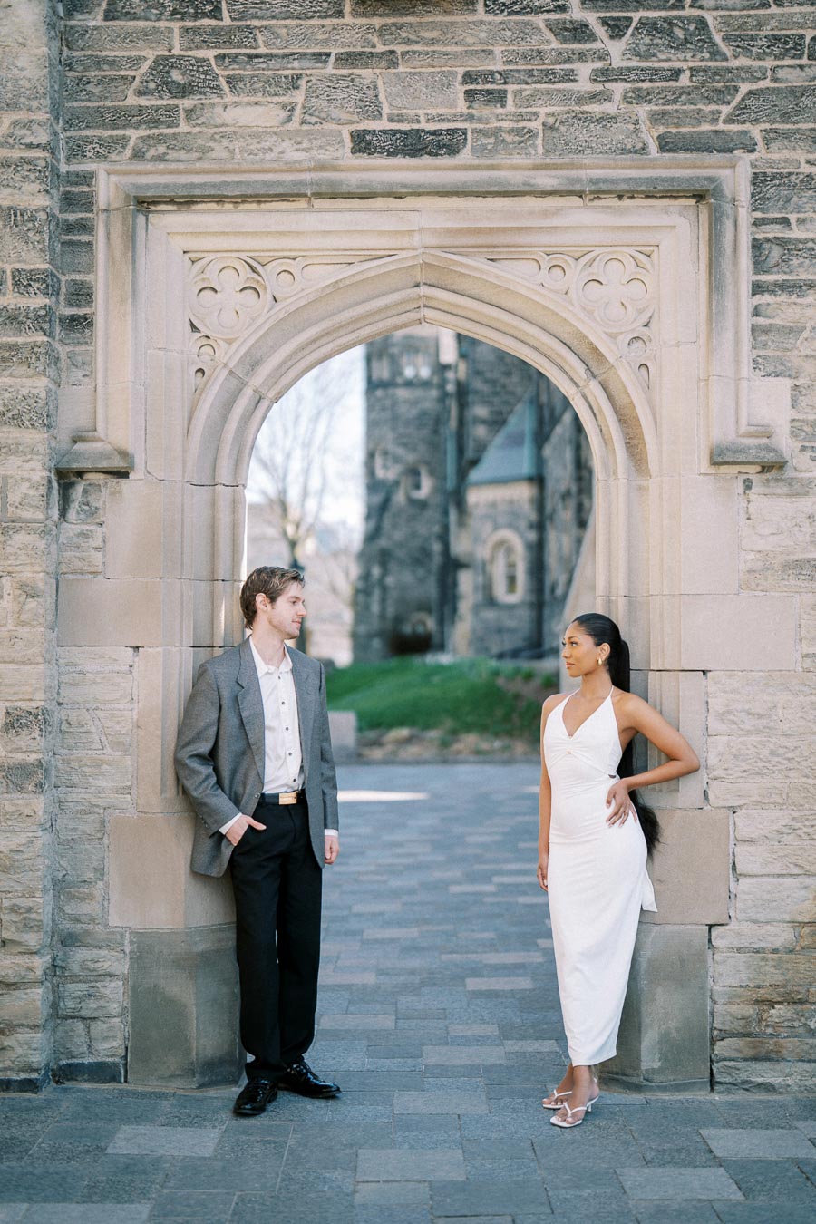 A man in a gray suit and a woman in a white dress stand in an ornate stone archway of a historic building, with a castle-like structure visible in the background.