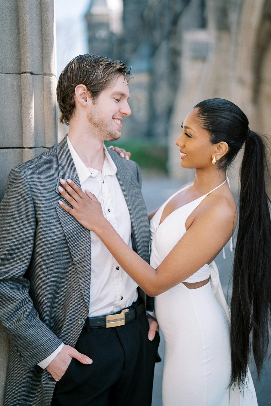 A couple standing closely, smiling at each other outside a stone building. The woman in a white dress has her hand on the man's suit jacket, capturing a romantic moment.