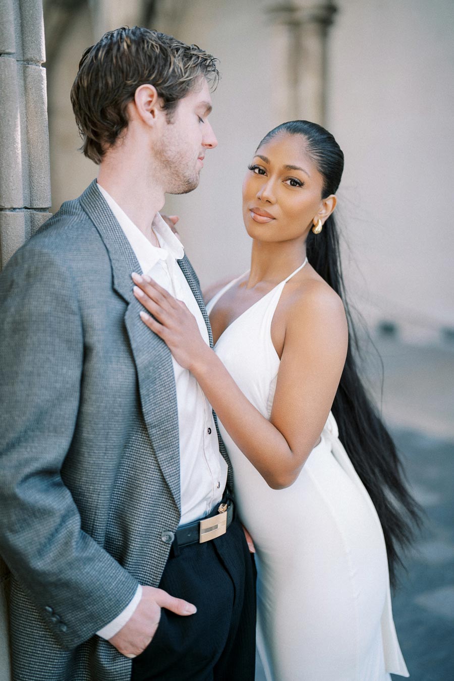 A stylish couple poses in elegant attire, with the woman in a white dress and the man in a gray suit jacket, standing close with a soft focus background.