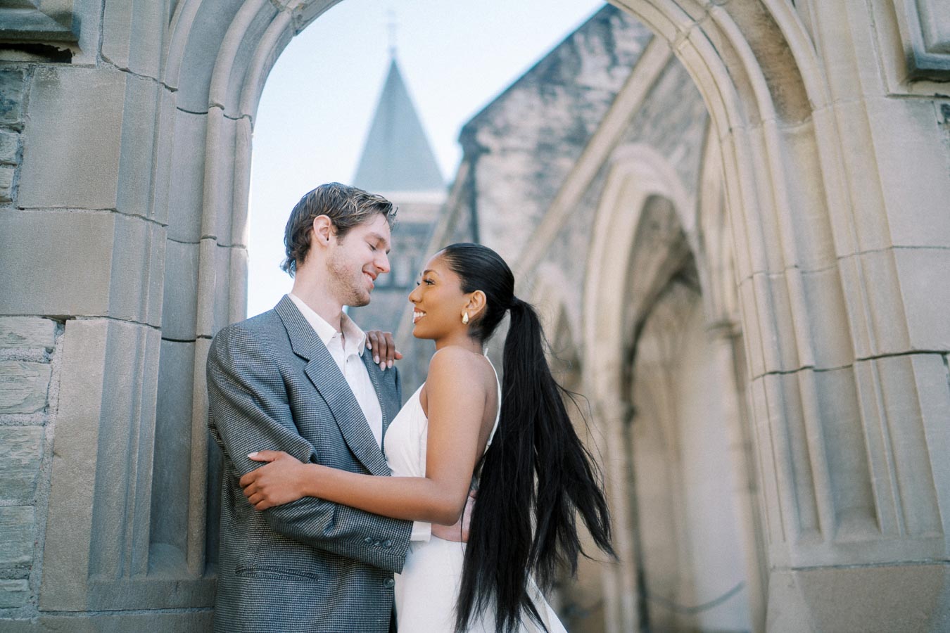 Smiling couple embracing under stone archway at historic building, showcasing a romantic and elegant atmosphere.