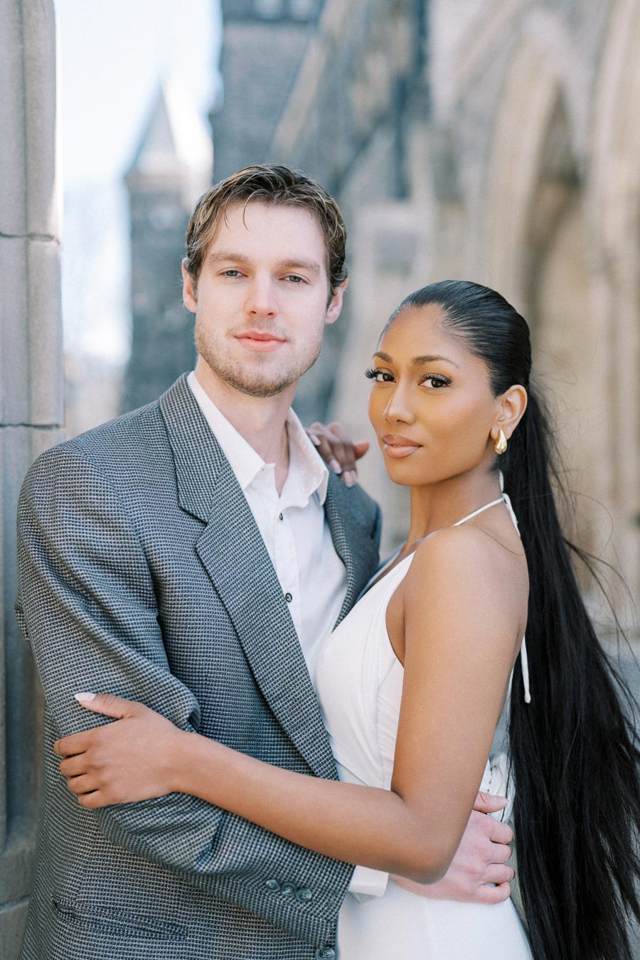 A couple posing closely in elegant attire outside a stone building, showcasing style and affection.