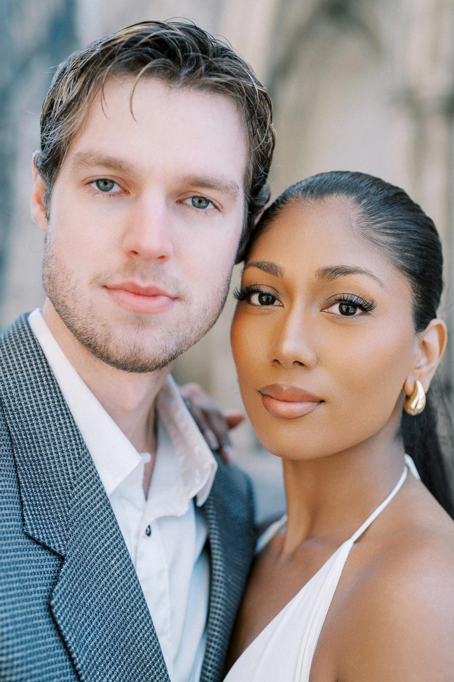 A couple posing closely in an outdoor setting, both dressed in formal attire. The young man is wearing a grey suit jacket and white shirt, while the young woman is in a stylish white halter dress, with her hair pulled back elegantly.