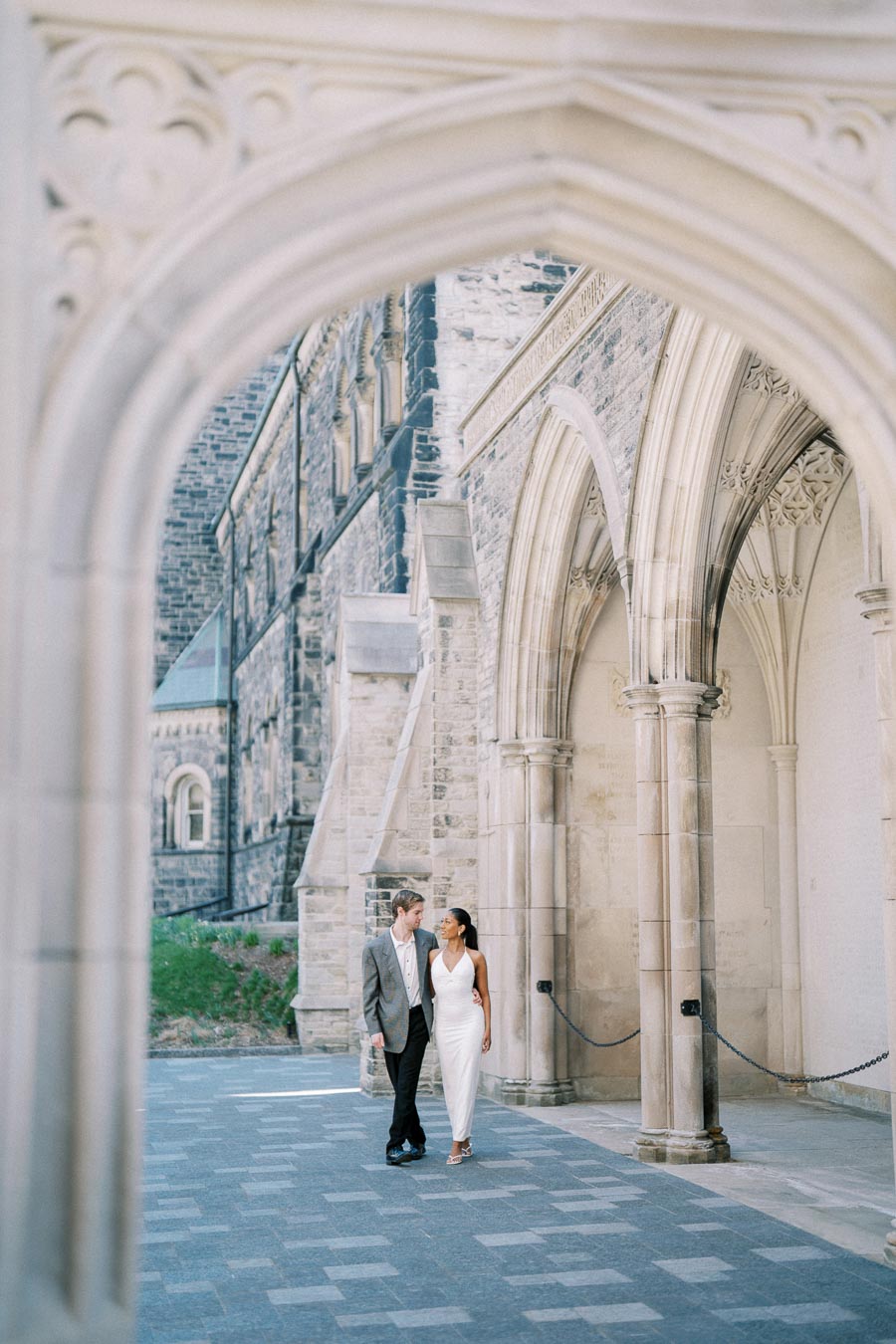 A couple walking arm in arm through a historic stone archway, dressed in formal attire, with an elegant architectural backdrop.