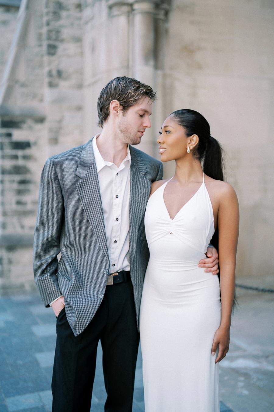 A couple standing close together outside a building, with the man wearing a gray suit jacket and the woman in a white dress, both looking at each other affectionately.