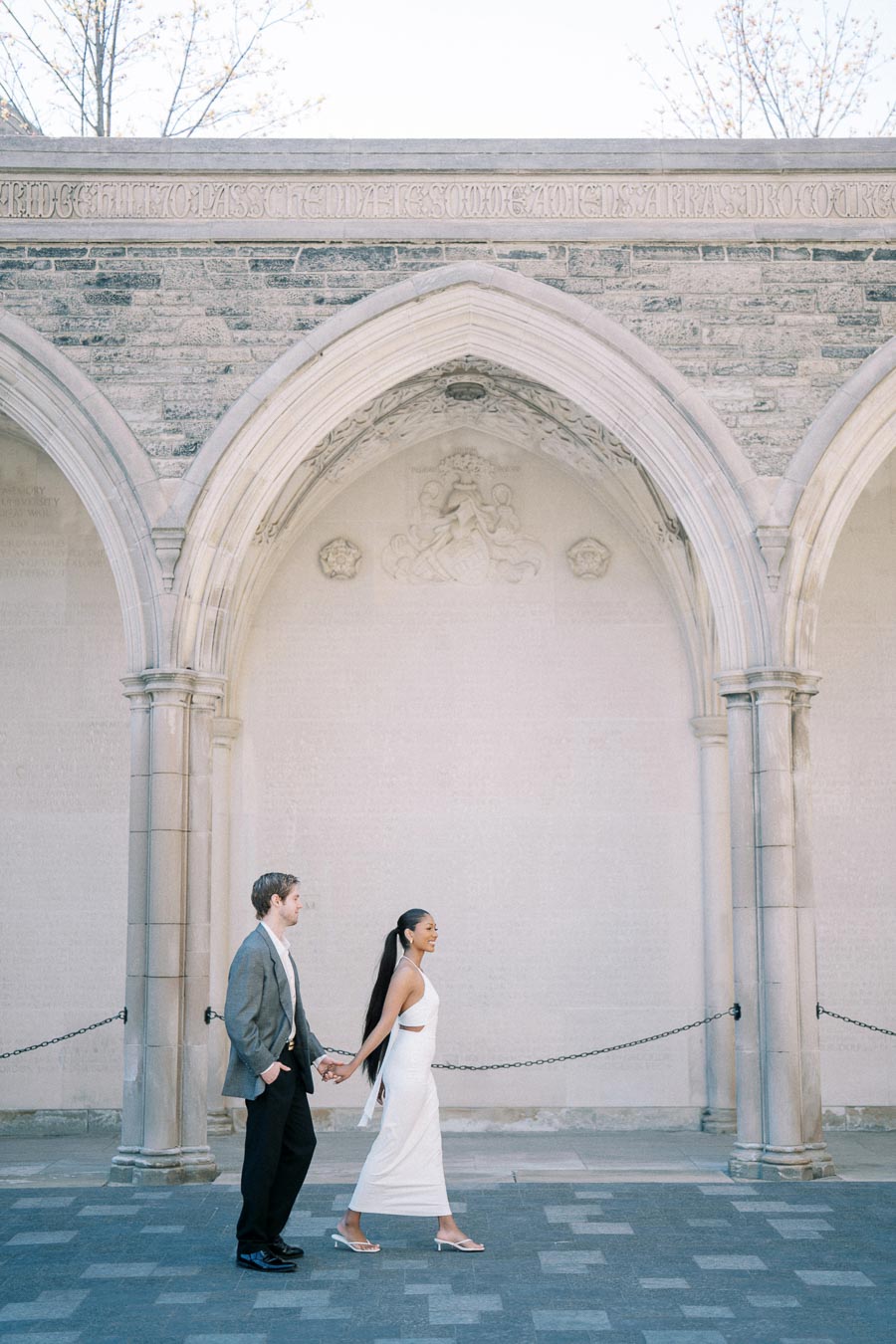 A couple holding hands while walking past a historic stone archway, showcasing elegant architecture and timeless romance.