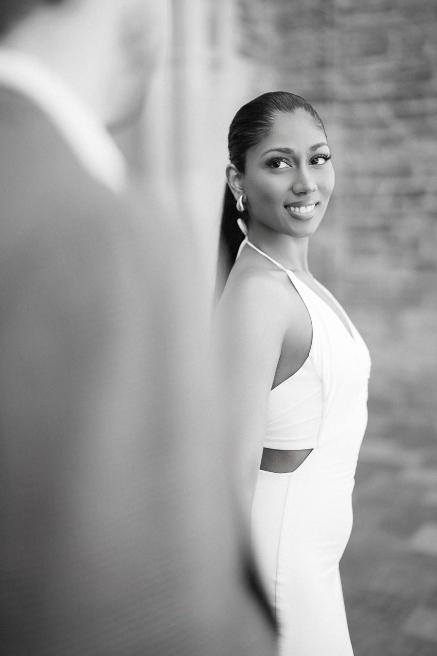 Black and white portrait of a woman in a stylish white dress smiling while looking over her shoulder, with a blurred figure in the foreground and a textured brick wall in the background