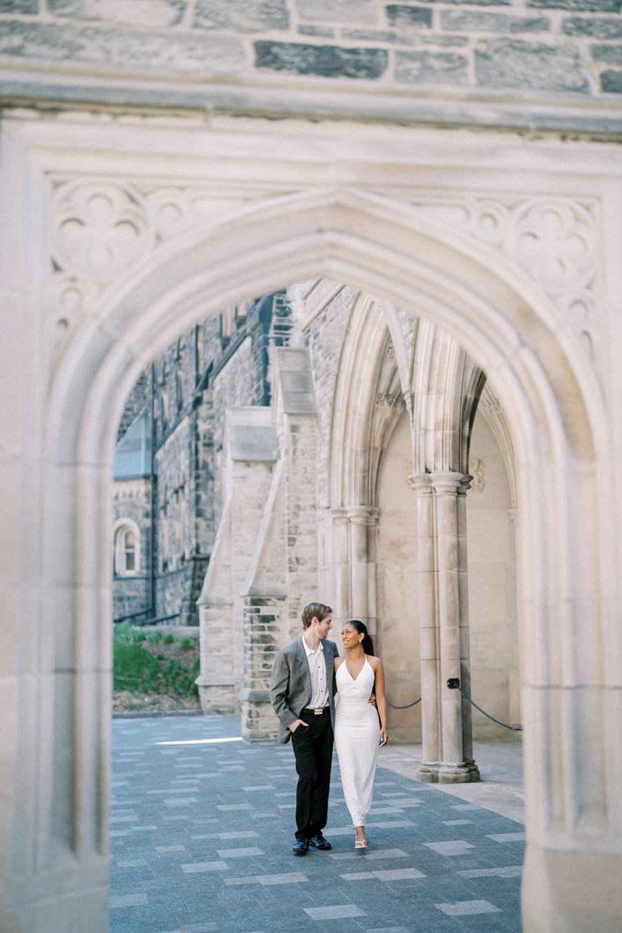 A couple walking arm in arm through a historic stone archway, wearing formal attire, with a picturesque old building in the background.