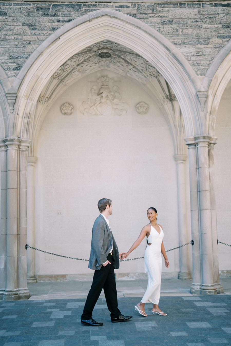 A couple walking hand in hand under elegant stone archways, with a woman in a white dress looking back at the man in a gray suit, set against a historic architectural backdrop.
