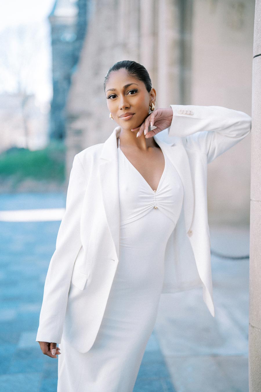 Elegant woman in a white suit leaning against a stone wall, exuding confidence in a serene urban setting.