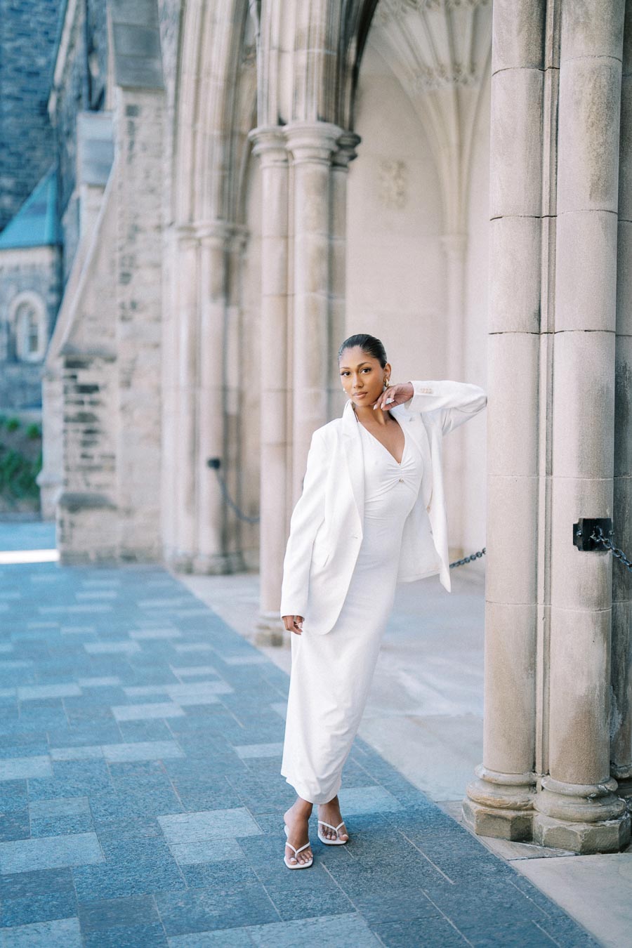 Elegant woman in a stylish white dress and blazer, posing confidently by a historic stone archway with detailed columns.