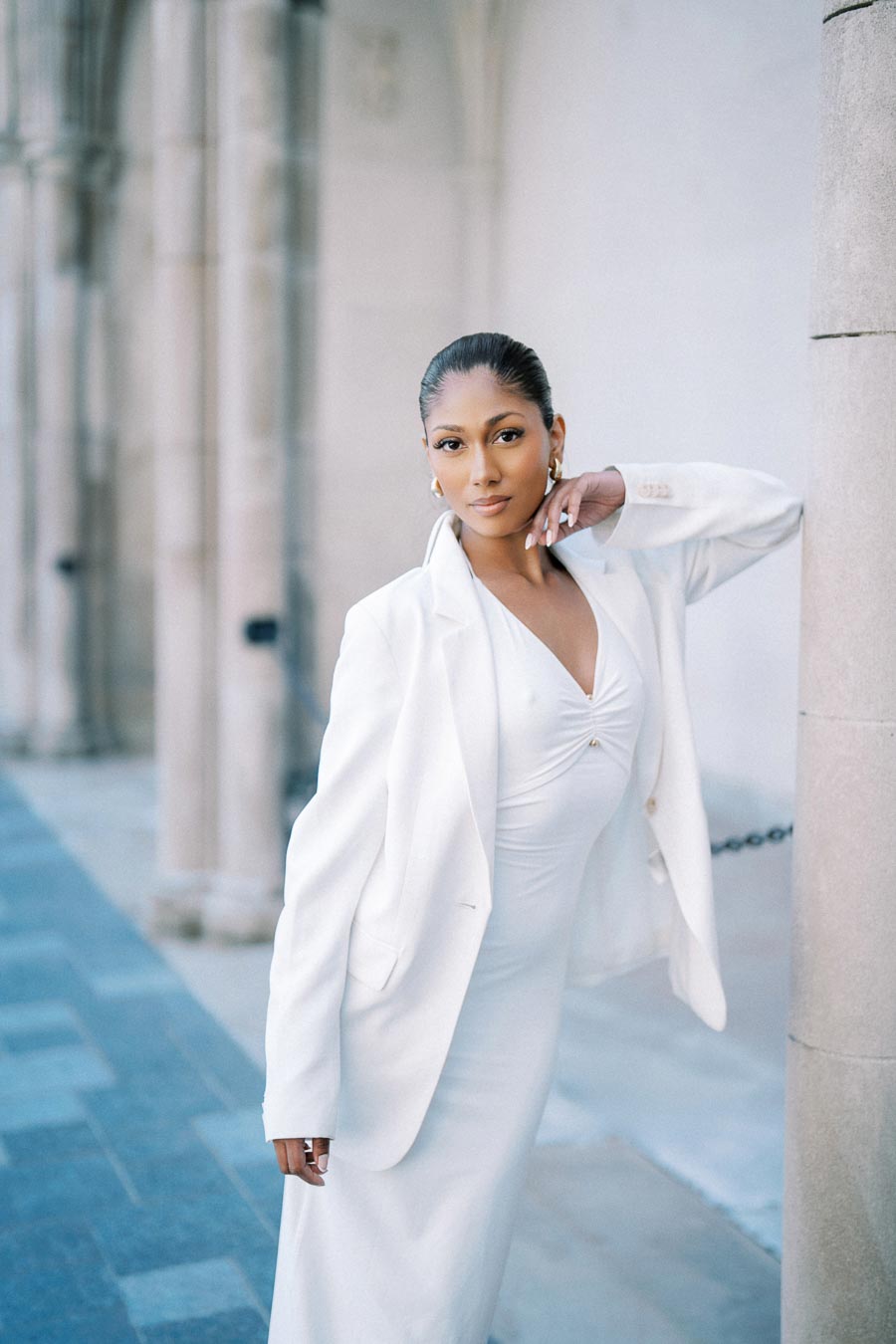A woman in a stylish white dress and blazer poses confidently in an elegant architectural setting with stone pillars.