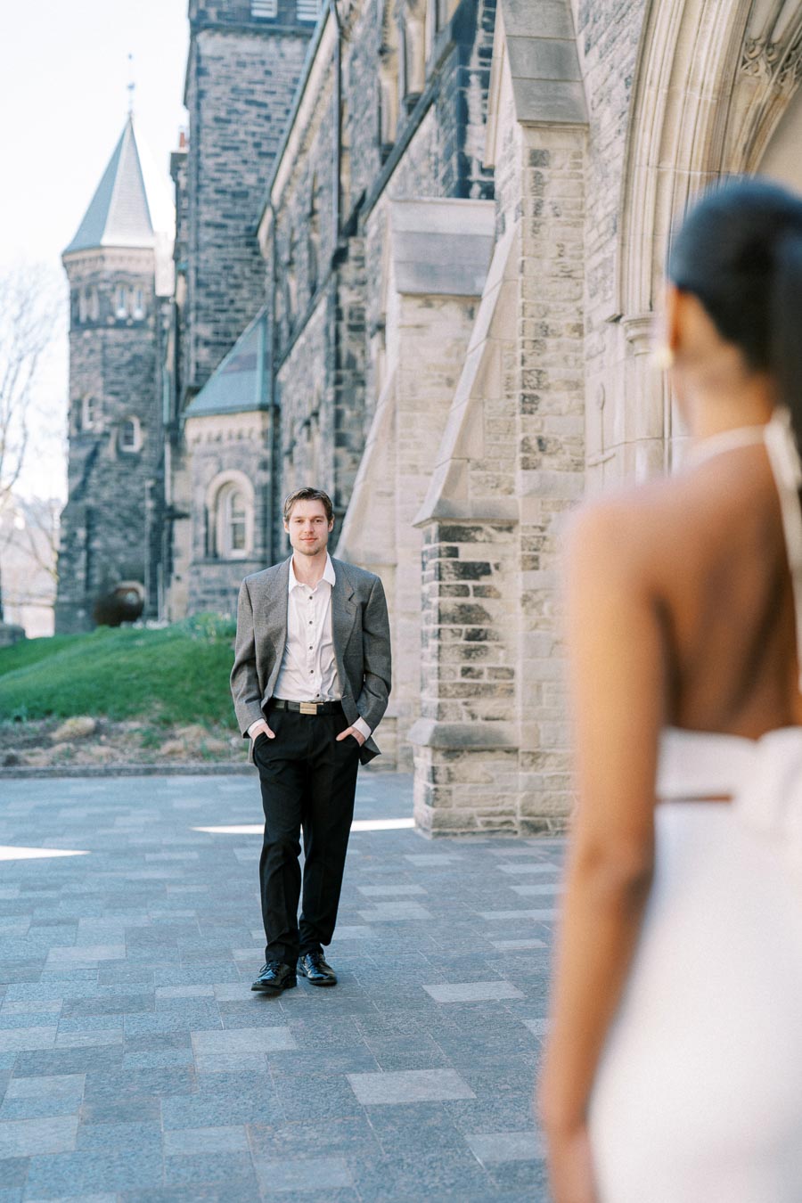 Man in a suit walking towards a woman in a white dress, with a historic stone building in the background.