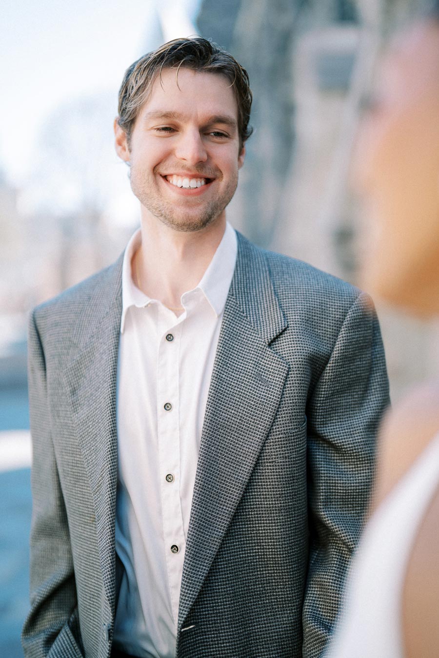 Man in a grey suit smiling outdoors with blurred background, wearing a white shirt, exuding confidence and professionalism.