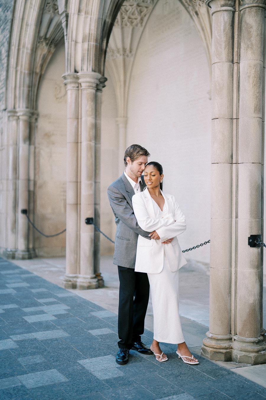 A couple embracing in an elegant, historic stone archway, with the woman wearing a white suit and the man in a gray blazer, conveying romance and elegance.