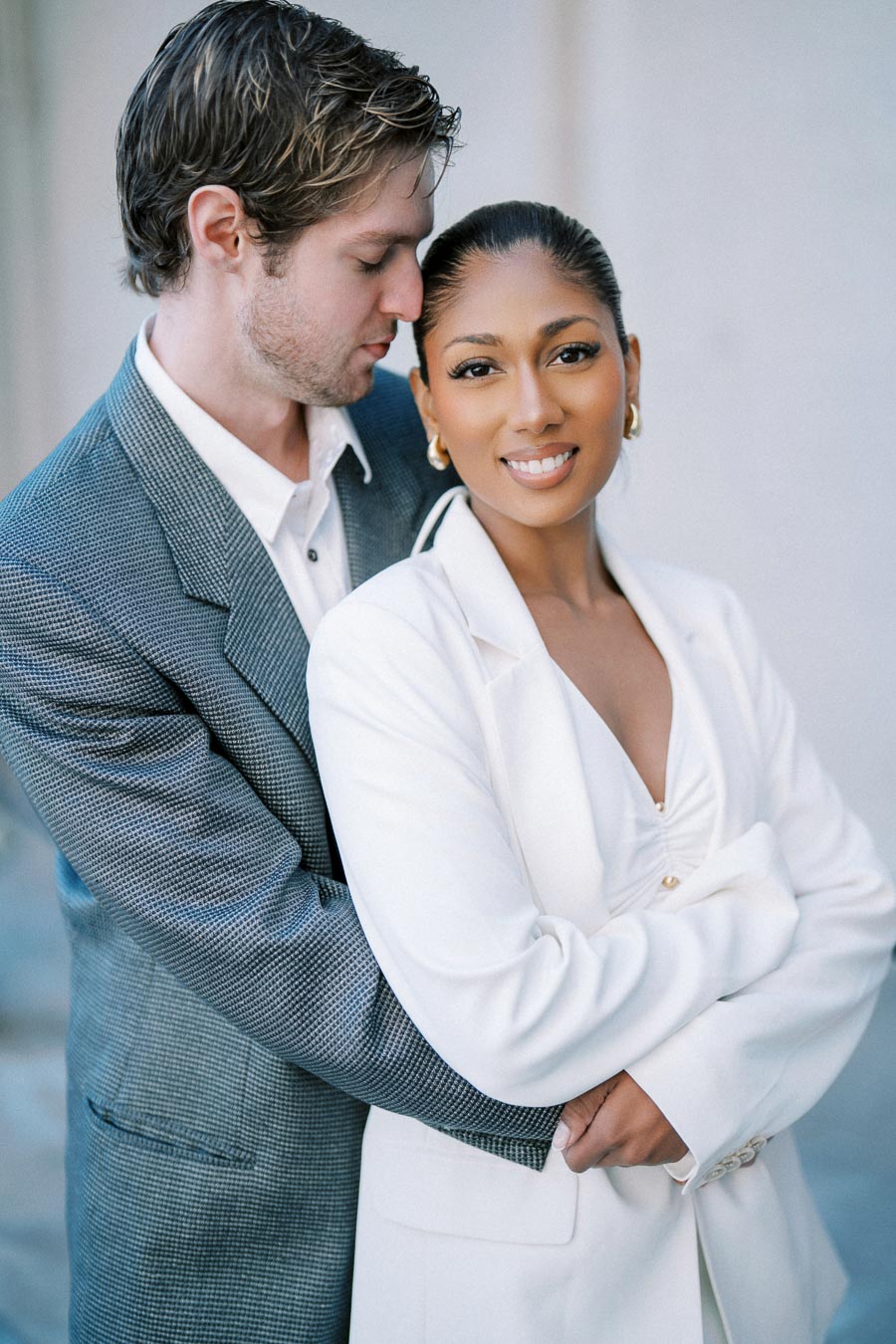 A smiling couple embracing, dressed in elegant white and gray suits, against a soft, neutral background.