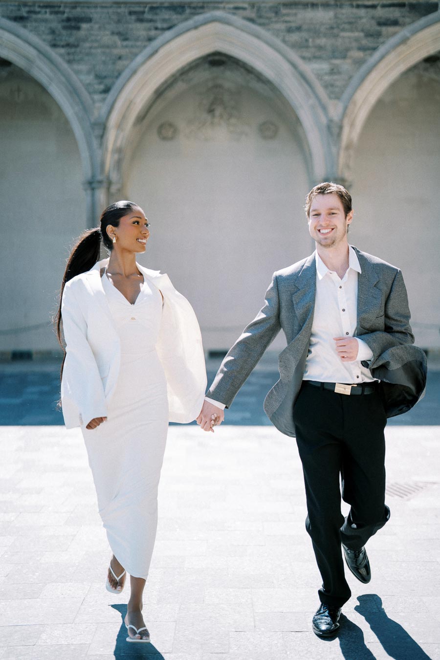 A couple holding hands while walking together at the University of Toronto, both dressed in formal attire.