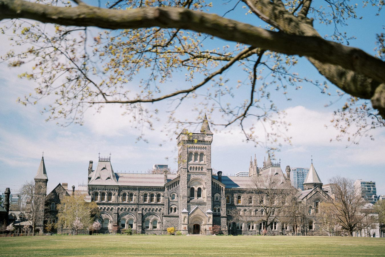 Alt text: Historic university building with Gothic architecture under a clear blue sky, framed by tree branches in the foreground.
