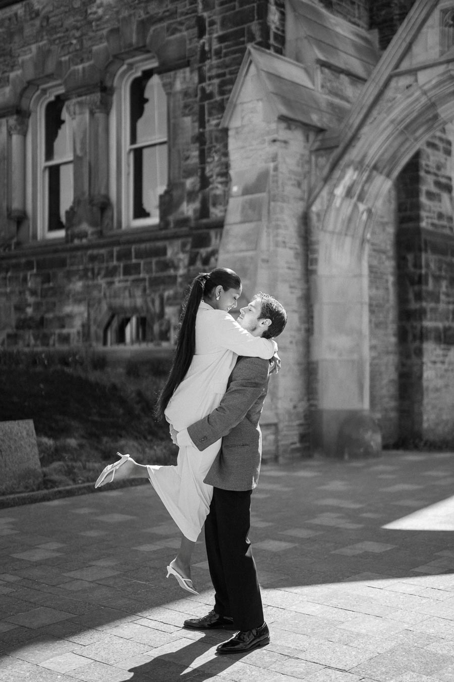 A black and white romantic photo of a couple embracing in front of an old stone building, with the woman in a dress and heels being lifted by the man in a suit, conveying joy and love.