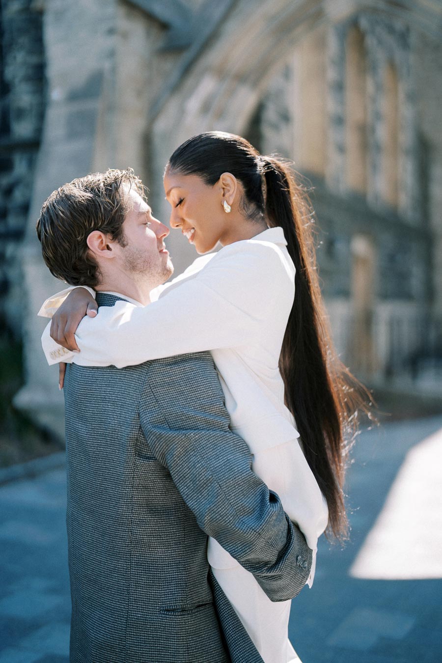 Happy couple embracing outdoors, enjoying a romantic moment in a sunlit setting with historic architecture in the background.