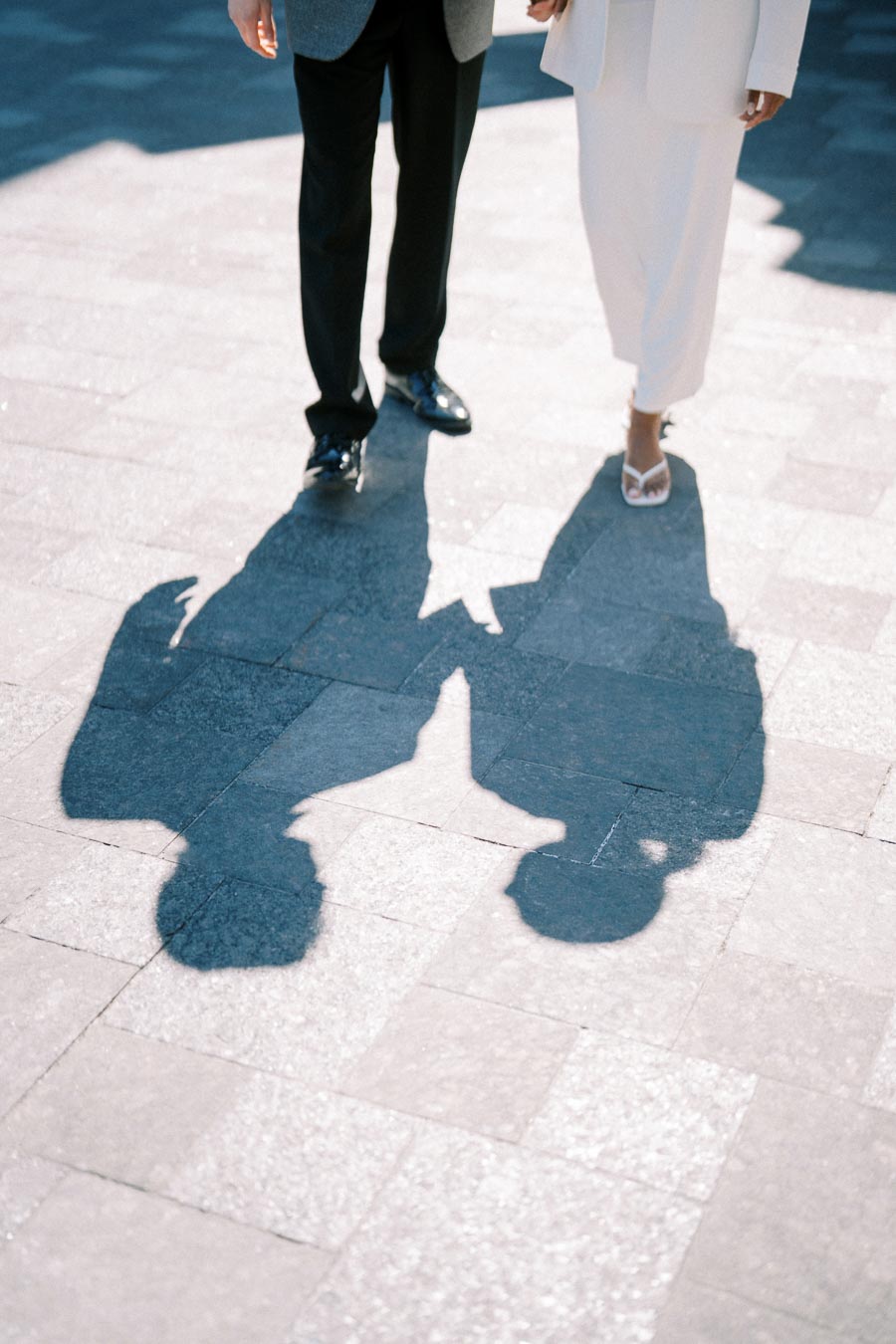 Two people walk hand in hand, casting shadows on a sunlit pavement.